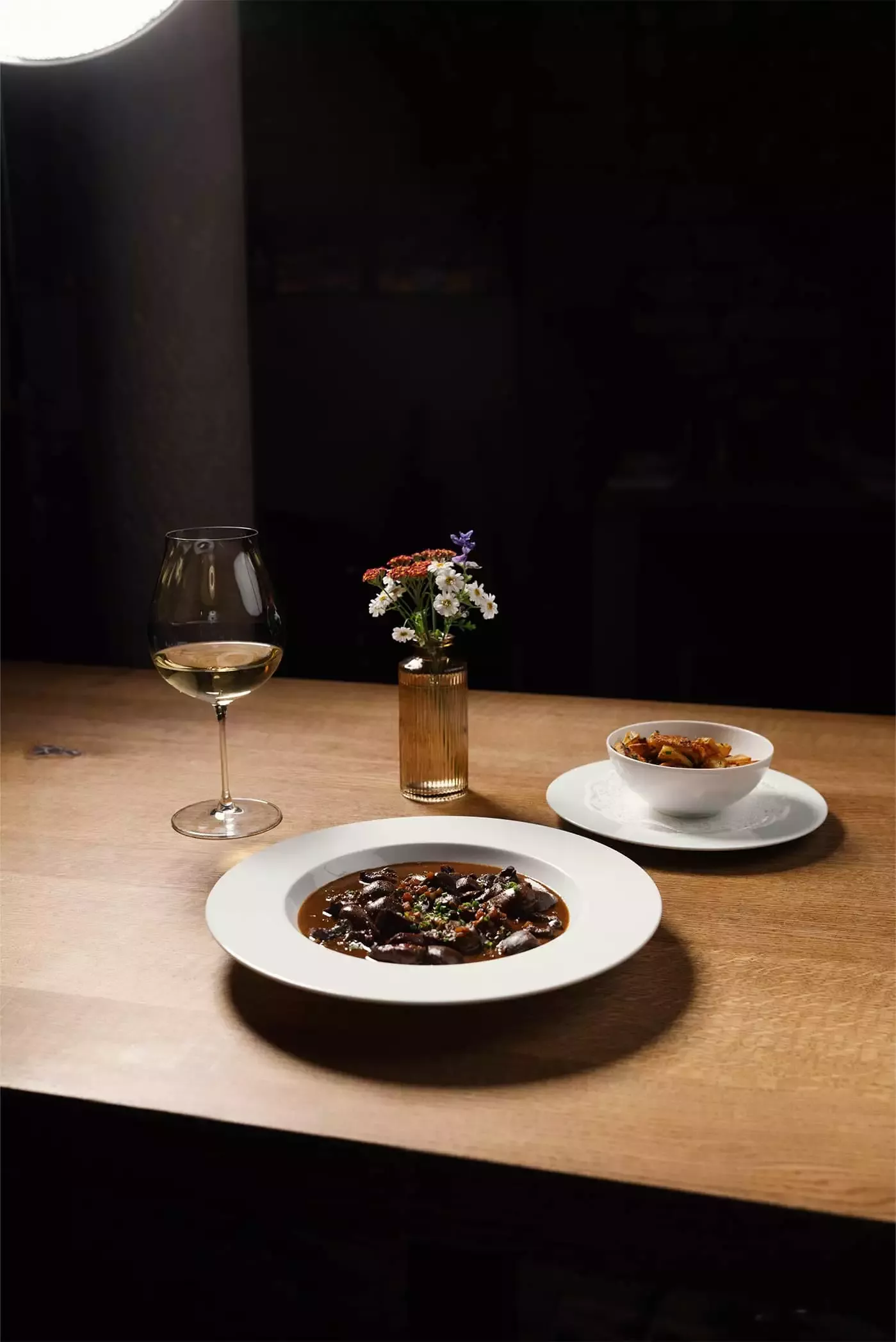 Ein gedeckter Tisch mit einem Teller reichhaltigem, dunklem Eintopf, garniert mit Kräutern, einer Schüssel mit gemischtem Gemüse, einem Glas Weißwein und einer kleinen Vase mit bunten Blumen. Der Hintergrund ist dunkel und hebt das Essen und das Dekor hervor.