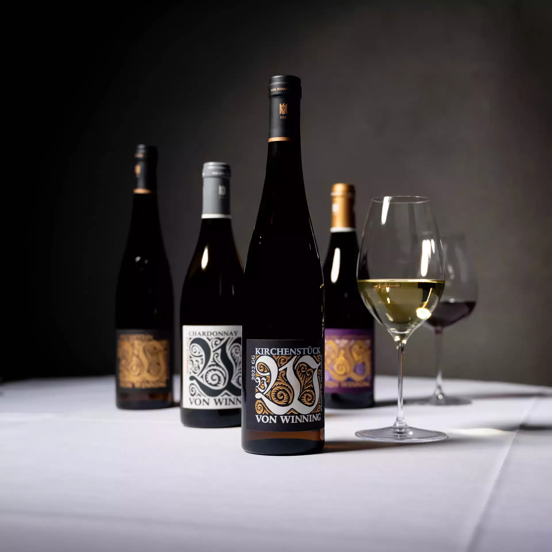 Vier Weinflaschen von von Winning auf weiß gedecktem Tisch, im Vordergrund eine Flasche Kirchenstück und ein Glas Weißwein.