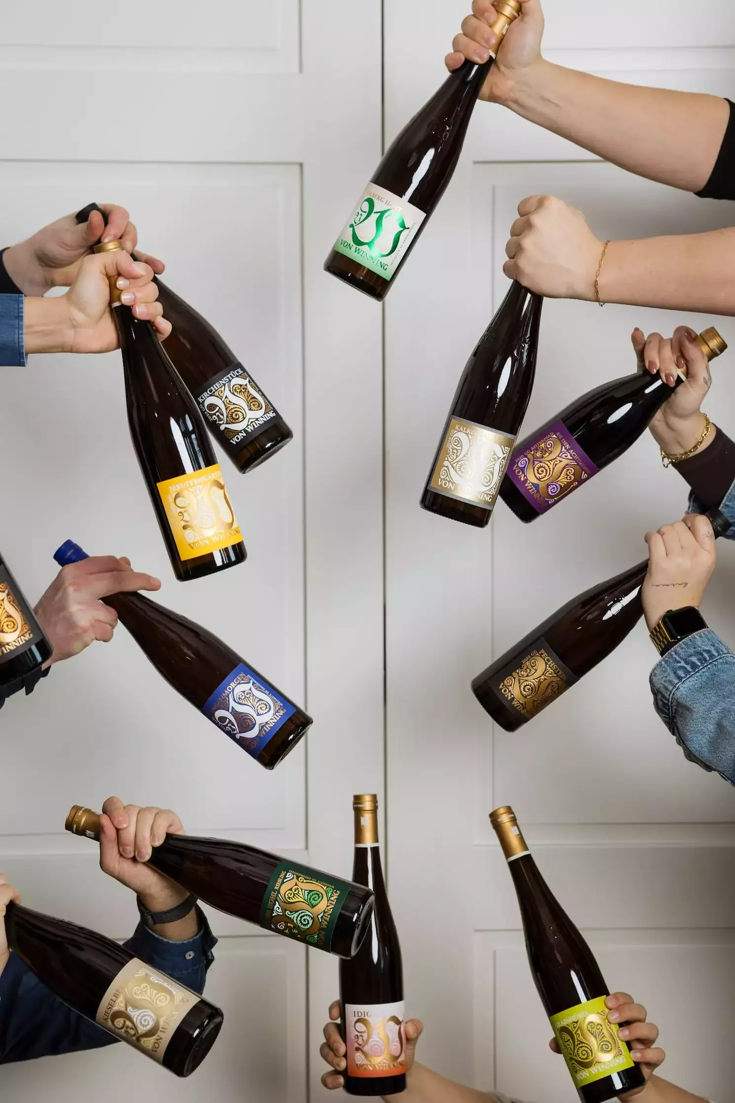 Mehrere Hände halten jeweils eine Weinflasche der Marke „Von Winning“ vor einem weißen, zweiteiligen Schrankhintergrund in die Bildmitte. Insgesamt sind 12 unterschiedliche Flaschen mit verschiedenfarbigen Etiketten zu sehen, darunter grün, gelb, blau, orange, lila und schwarz. Die Flaschenetiketten zeigen ein auffälliges, geschwungenes Ornamentdesign mit der Aufschrift „Von Winning“. Jede Flasche wird von einer Person gehalten, jedoch sind nur die Hände und Unterarme sichtbar, manche mit Armbändern oder Uhren. Die Flaschen sind in einem dynamischen, fast kreisförmigen Muster um die Bildmitte herum angeordnet, was dem Bild eine lebendige, feierliche Stimmung verleiht.